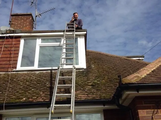 Roofer at the top of a ladder on a dormer, above a tiled roof covered in moss