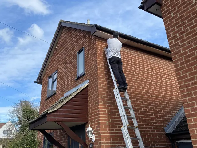 Roofer up a ladder working on the fascias and guttering of a red brick house